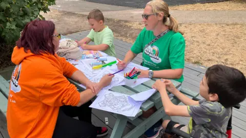 Shariqua Ahmed/BBC Two women and two children sitting on a green bench, doing colouring.
