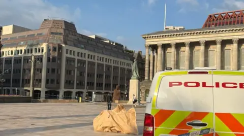 A police vehicle is on the right, with Birmingham Town Hall in the background. Another building is on the left.