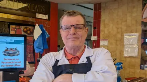 Robert Price is stood in front of the counter in his shop. He wears dark rimmed glasses, a white shirt and a blue apron.