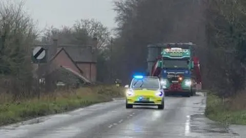 Suffolk Highways A yellow and white police car, with blue lights shining, driving along the middle of a road in front of a blue and green lorry