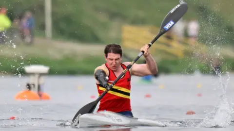 Jon White A man with brown hair in a red top in a small white kayak with a black paddle. One of his arms is missing and he has a black prosthetic.