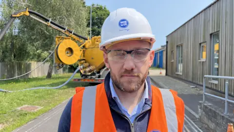 George Carden/BBC Nick Mills who works for Southern Water wearing an orange high vis which says Water for Life on it. He is also wearing a white hard hat and behind him is a Southern Water tanker.