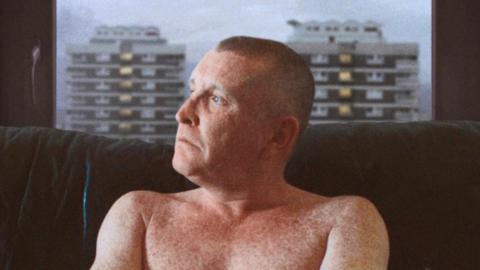 Joe McNally in The Flats pictured topless on sofa with dog and New Lodge flats visible through window behind him