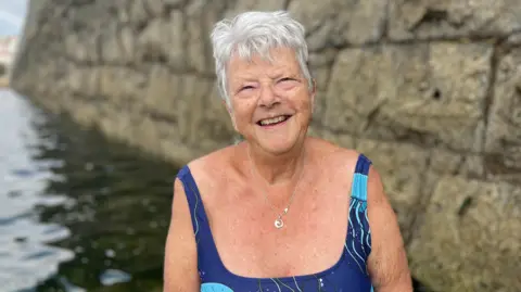 A woman with grey hair looking towards the camera, she's wearing a Patois jewelery necklace and a blue swimsuit.