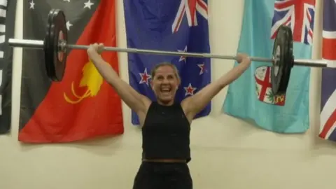 Dawn pictured holding a large dumbbell over her head and smiling widely. She's wearing black leggings, a black sleeveless top and white and pink trainers, and has her blonde hair tied up in a ponytail. She's photographed indoors, inside her gym - which has cream walls and a brown wooden floor. Behind her are multiple flags, including the Union Flag, hung up on the wall.