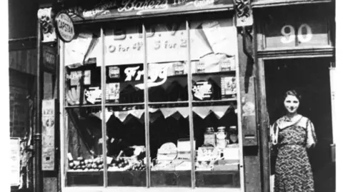 Manchester Jewish Museum A young woman in a dress stands in the doorway of a shop. The window has products laid out and the doorway reads the number '90'.