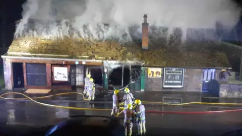 Kelly's Cafe A row of single storey buildings on fire. Smoke coming from the roof, with firefighters using hoses to stem the blaze. 