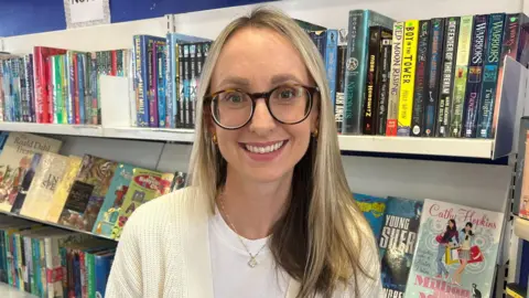 Laura Parry mid-length hair, she wears a white top and cardigan, and round dark glasses. She stands in front of a bookshelf which is full of colourful books.