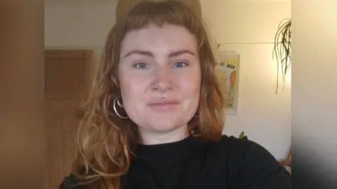 Ruby Love Love wearing a black top and her long, wavy strawberry blonde hair down with a fringe. She is wearing silver hoop earrings and face piercings and is smiling lightly at the camera.