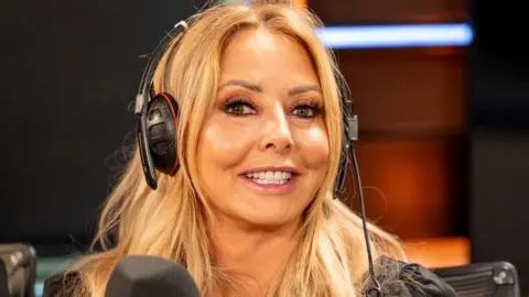Carol Vorderman pictured in a BBC radio studio. She has long blonde hair and is smiling at the camera. She is wearing headphones, whilst sat in front of a microphone.