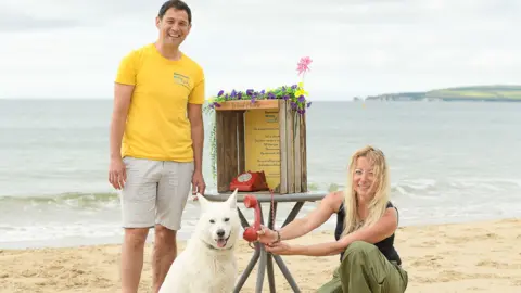 Heather Field A man and a woman using a red wind phone on a table at a beach on a cloudy day. They are smiling. A white dog is beside them. The woman is pointing the receiver to the dog. The instructions of how to use the phone are written on the box it is placed in. There are yellow and purple morning glories and a pinwheel on top of it. The sea is seen behind them.