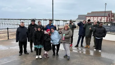 A group students standing in rows in front of a pier. They are wrapped in warm coats and it looks like it has been raining. Some of the students are saluting at the camera.