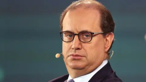 Mark Wiseman, seen in 2023 and from the shoulders up, wears dark rimmed glasses and a dark suit and patterned tie. 