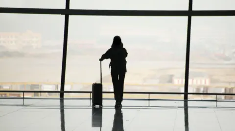 Getty Images The silhouette of a person with a suitcase standing at a big glass window at an airport 