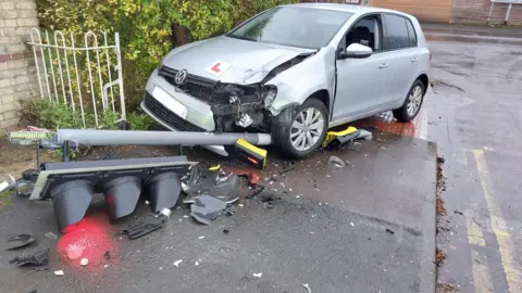 Cambridgeshire Constabulary A silver car has mounted a pavement and is missing its front headlight. Underneath the car is a pole with a traffic light, now smashed, on the end. On the front of the car is an L plate.