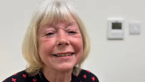 Annette Murray-Hamilton. She has blond hair in a bob and green eyes. She is wearing a black blouse with a red cross pattern and she is smiling at the camera. She is inside a room with white walls and the background is blurred.