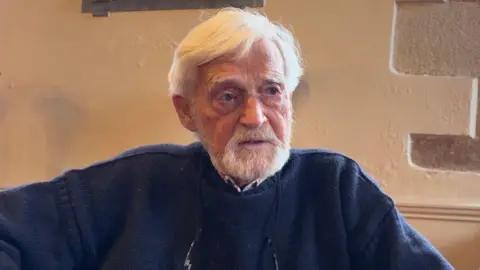 A man sits in front of a white stone wall. He has a white beard and white hair. He is wearing a navy blue jumper with a shirt underneath.