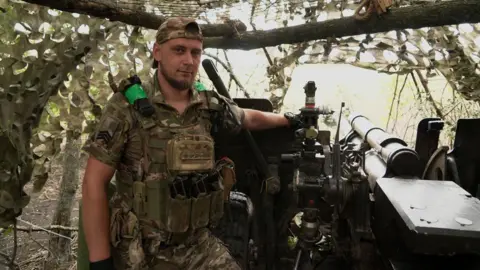 Ukrainian commander Dmytro, of the 15th Brigade National Guard, stands next to a weapon wearing military fatigues.