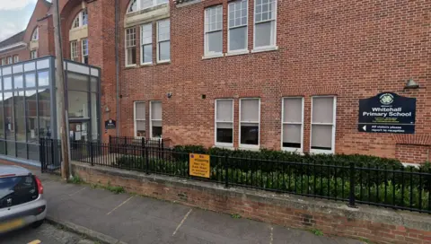 A red brick primary school with a glass reception area. A sign reads Whitehall Primary School. A car is parked on the street outside. 