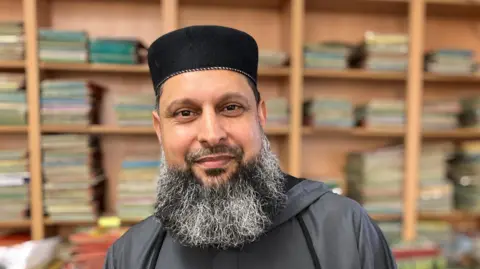 Najabat Ramzan has a bushy beard which is speckled with grey and a neat moustache. He is wearing a brimless hat and a grey top. Behind him you can see lots of bookcases. 