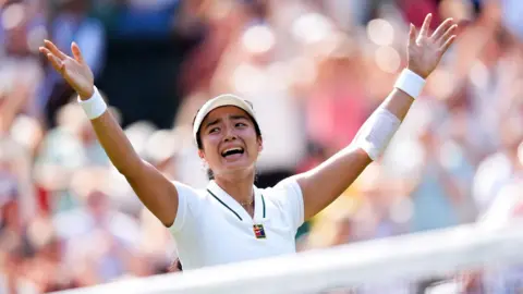 PA Media Filipina tennis player Alexandra Eala raises his arms after a Wimbledon match, net in front of her and a blurred image of the crowd in the background.