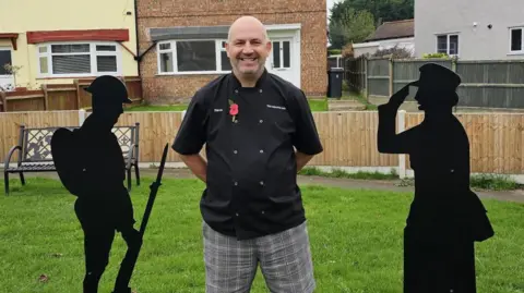 Michael Keohan/BBC Dave Hubbard stands next to the two statues on the grass. He wears a chef's tunic and gray checkered trousers