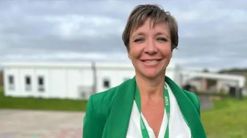 Stuart Woodward/BBC Jayne Crowley wears a green jacket with white blouse underneath, and a green staff lanyard around her neck. She has short cropped brown hair and is smiling at the camera. She stands in front of a white building with a patch of grass in front of it, next to a school playground.