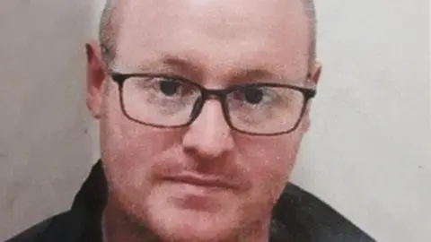 Police handout Matthew Armstrong is looking at the camera, wearing glasses. He is balding, and has a faint, stubbly beard. He is wearing a coat.