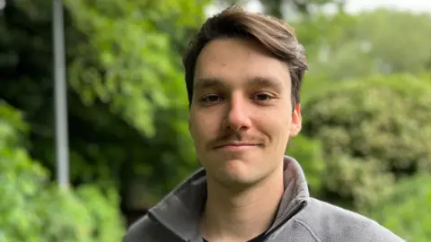 Francisco Campis is standing off centre to the right of the frame. He has short brown hair and moustache and is wearing a black T-shirt and a grey jacket. He is standing in front of a row of trees and bushes.