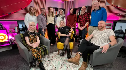 The podcast team posing for a group photo. There are six women, including Danni Diston and Sarah Buck and three men, including Edward Rowe and Mark Jenkin. The studio is brightly lit with pink and purple neon.