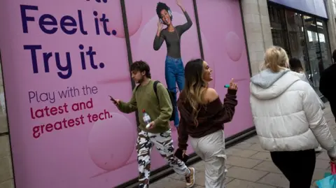 People walk past a Currys shop with a pink sign in the window saying "Feel it. Try it. Play with the latest and greatest tech."