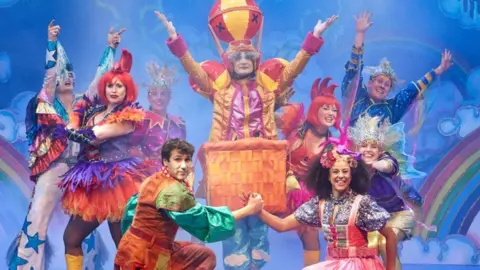 Pantomime actors dance on stage while wearing colourful outfits.