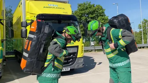 Two men are wearing green and yellow uniforms and green helmets. They are putting black backpacks onto their backs.