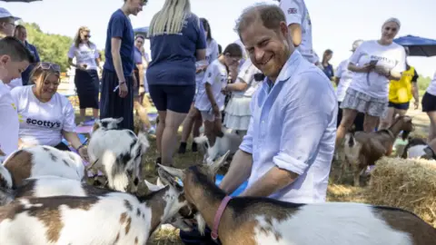 PA Media Prince Harry, wearing a light blue shirt, smiles at the camera while sitting down surrounded by goats.