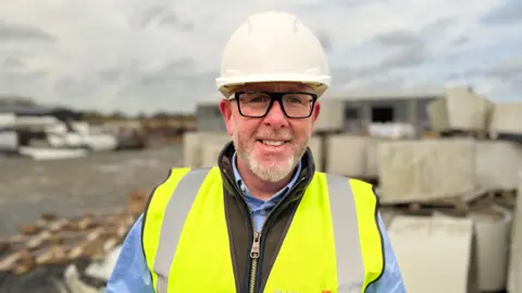 Kieran Kelly, a man with black square-framed glasses and a grey beard, smiles at the camera in an industrial yard. He is wearing a white helmet, a yellow high-viz vest, a blue shirt and a brown body warmer. 