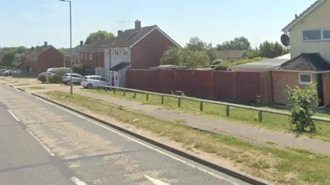 A view of Whitmore Way. There are houses to one side and a pathway with scrubby grass and a wooden fence