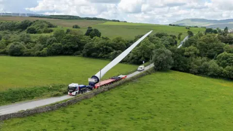 CWP Energy A blade lifter carrying a wind turbine blade through a rural landscape with a small escort van following behind