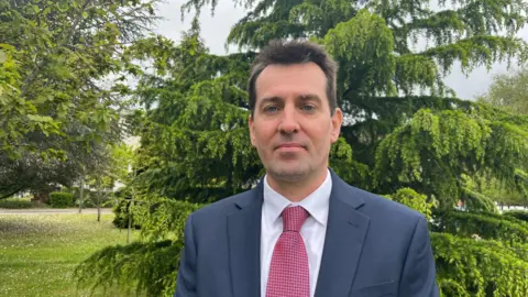 A man with dark brown hair wearing a blue jacket, a white shirt and a pink tie standing in front of a tree