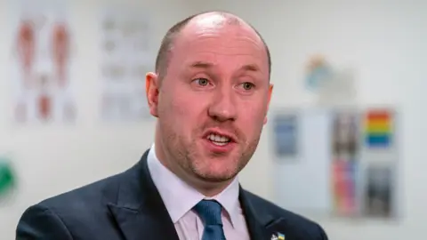 PA Media Neil Gray looking slightly off camera. He is wearing a dark suit over a white shirt with a dark blue tie.