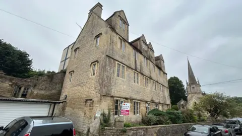 BBC Three story Cotswold stone apartment block with church in background