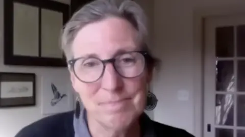 Martha Williams, a woman with grey tied back hair, wearing large round rimmed glasses, and large earrings. She is sat in a white room with pictures on the wall, including a drawing of a flying bird