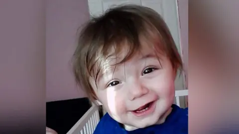 Family photo Ethan Ives-Griffiths, a small toddler with messy brown hair and dark brown eyes, half smiling at the camera. He looks like he is in cot and is wearing a blue top