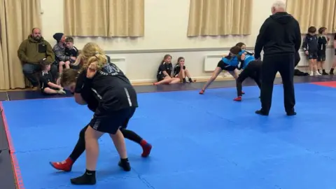 Ollie Rawlinson/BBC Two pairs of girls wrestling on a blue padded mat. They each have each other in a hold. There are people sitting around the outside of the mat meanwhile two instructors watch both pairs closely.