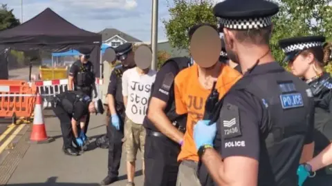 Just Stop Oil protesters wearing branded tops being arrested by police while standing on the street.