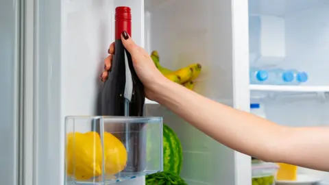 Getty A hand placing a dark glass bottle with a red screw cap into an open refrigerator. In the foreground, two yellow lemons sit in a clear door shelf compartment. Inside the fridge, a watermelon, a bunch of bananas, several plastic water bottles, a lidded food container and a block of cheese are visible on shelves. The refrigerator interior is white and brightly lit.