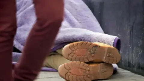 Pair of feet with boots under a purple blanket on the ground in an outdoor area. A blurred pair of legs walk past in the vicinity. 