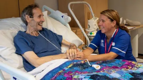 A hospice patient with a nasal drip talks with a nurse in blue uniform who is holding his hand.