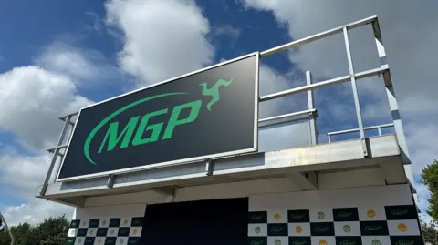 BBC The Manx Grand Prix podium, which is a platform with railing around it and a large board featuring the green MGP logo on the front. The sky above is blue with white fluffy clouds.