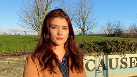 Cara Hunter looking ‌at the camera with‌ a neutral ⁤expression.‌ she is wearing⁢ a brown coat. Behind her a⁢ grassy ⁢hill and a few trees.