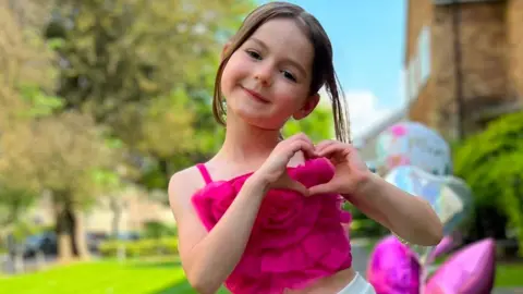 Leonna wears a purple floral crop top and is making a heart symbol with her hands. She has brown hair and smiles. She is outside and there is grass and trees behind her.